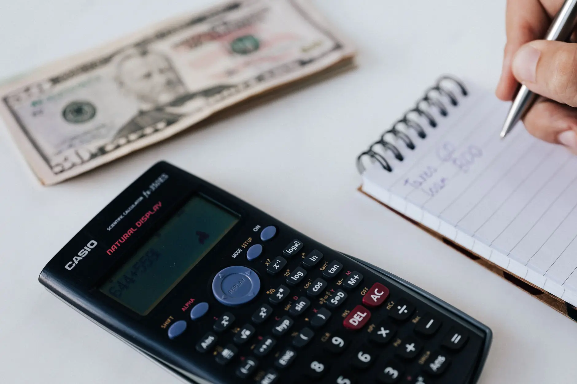 Mano haciendo cálculos junto a un cuaderno y un billete de dólar