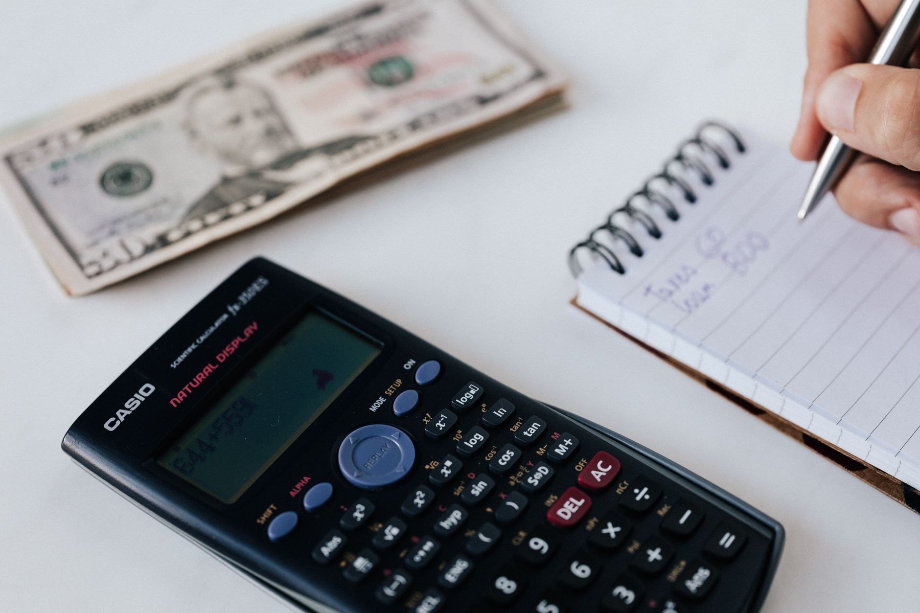 Mano haciendo cálculos junto a un cuaderno y un billete de dólar
